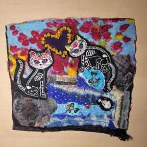 May include: A colorful fabric art piece featuring two embroidered skeleton cats with a heart and fish designs. The background is a patchwork of blue, red, and black fabrics.