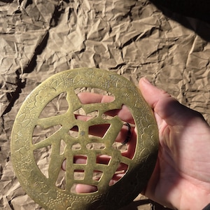 May include: A round, antique brass object with a central Chinese character design. The outer edge features a floral pattern. The object is held in a hand against a textured, brown paper background. The brass has a warm, aged patina.