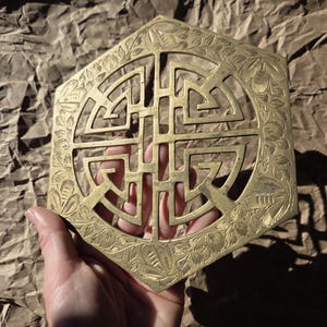 Puede incluir: Un salvamanteles hexagonal de latón con un intrincado diseño calado. El centro presenta un patrón geométrico, rodeado por un borde floral. El salvamanteles se sostiene sobre un fondo marrón texturizado.