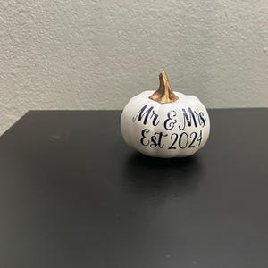 May include: A white decorative pumpkin featuring a metallic gold stem and black cursive text on the front that reads Mr & Mrs Est 2024. The pumpkin has a smooth, rounded shape with subtle vertical ridges and a flat base for stable placement.