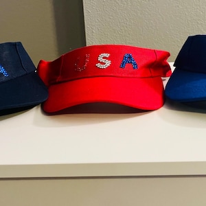 May include: Three visors in red, navy blue, and royal blue, each with the word "USA" spelled out in rhinestones. The visors are displayed on a white surface, showcasing their patriotic design.
