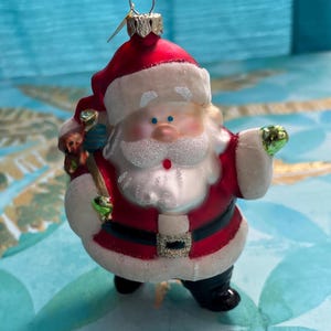 May include: A festive Christmas ornament shaped like Santa Claus. The ornament features a red suit with white trim, a black belt, and black boots. Santa is holding a green object and a sack of toys. The ornament has a glittery beard and a smiling face.