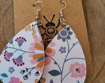 May include: Teardrop-shaped earrings with floral patterns. The earrings feature a white background with pink, purple, and blue flowers and green leaves. The earrings are hanging from silver hooks. The background includes a brown tag with the text "Just Beeutiful".