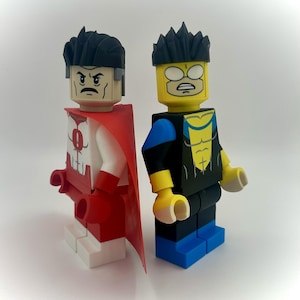 May include: Two custom-made action figures, inspired by the character Invincible, stand side-by-side. One figure is in a red and white superhero suit with a red cape. The other figure is in a black and blue suit with yellow accents. Both figures have black hair.