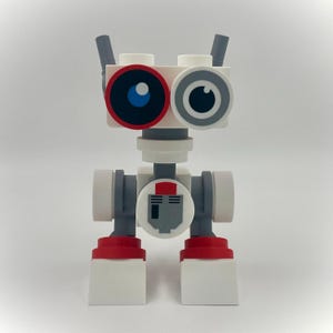 Könnte beinhalten: Ein Roboter-Spielzeug aus weißen, grauen und roten Bausteinen. Er hat zwei Augen, eines mit einer blauen Iris und rotem Rand, das andere mit grauem Rand. Der Körper hat ein kreisförmiges Design mit einer roten und grauen Grafik.