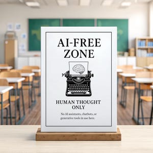 Op de afbeelding: Een wit bord met zwarte tekst met de tekst "AI-FREE ZONE" en "HUMAN THOUGHT ONLY" in een klaslokaal. Het bord toont een typemachine met een hersenillustratie. Het bord staat op een houten standaard.