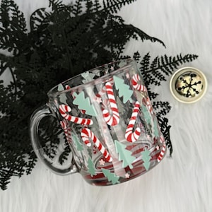 May include: A clear glass mug with a handle, decorated with red and white candy canes and green Christmas trees. The mug is on a white surface with a sprig of dark green foliage and a small gold bell.