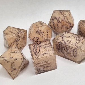 Pode incluir: Uma coleção de dados geométricos com designs de mapas vintage. Os dados têm várias formas, incluindo cubos, pirâmides e formas multifacetadas, com um padrão de mapa em tons de sépia visível em cada face. Os dados estão sobre um fundo branco.