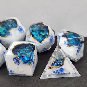 Puede incluir: Un conjunto de dados de resina hechos a mano en varias formas, incluyendo una pirámide y un cubo. Los dados presentan una base blanca con patrones florales azules y detalles dorados, rellenos de resina azul y verde brillante.