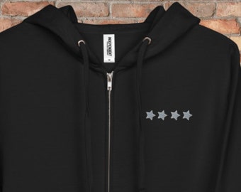 May include: Black zip-up hoodie with a drawstring hood and front pockets. The hoodie features three small, gray embroidered stars on the upper left chest. The zipper is silver. The hoodie is displayed against a brick wall.