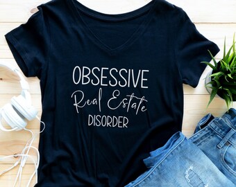 May include: Black v-neck t-shirt with the text "OBSESSIVE Real Estate DISORDER" in white lettering. The shirt is laid flat on a light wood surface with a pair of blue jeans and a white pair of headphones.