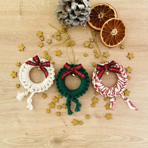 Op de afbeelding: Drie handgemaakte kerstkransen met rood-wit geruite linten en bellen. De kransen zijn gemaakt van wit, groen en rood garen en hangen aan een gouden touwtje.