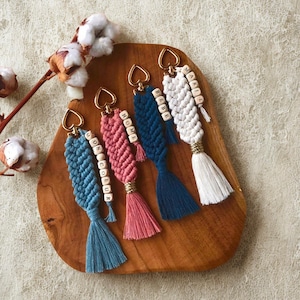 Peut inclure: Quatre porte-clés en macramé bleus, roses, marine et blancs, chacun avec un fermoir en forme de cœur doré et un pompon. Les porte-clés comportent des perles de bois épelant des mots comme "MON COEUR", "HOME", "BEBE" et "MARIE".