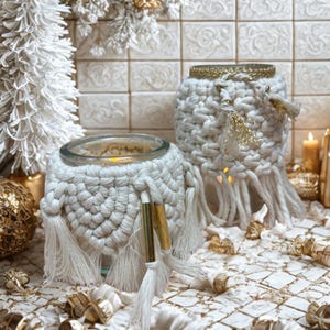 Peut inclure: Deux photophores en macramé blanc avec inserts en verre. Les supports présentent un design tissé avec des franges et des accents dorés. L'un est légèrement plus grand que l'autre. Le fond comprend un sapin de Noël blanc et d'autres éléments décoratifs.