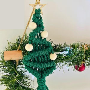 Può includere: Un ornamento natalizio fatto a mano in macramè, di colore verde scuro, adornato con quattro perle di legno chiaro e una stella di legno. L'albero è sospeso da un cordoncino di spago naturale, creando una decorazione festosa.