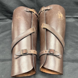May include: Pair of brown leather leg armor with buckle straps. The armor is cylindrical in shape, with a slightly flared top and bottom. The leather has a worn appearance, with some scratches and discoloration.