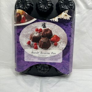 Puede incluir: Molde para brownie Bundt negro Nordic Ware Pro Cast con 12 moldes individuales. El molde está en una caja con fondo morado y una imagen de pasteles Bundt de chocolate con azúcar en polvo y frambuesas.