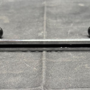 May include: A silver metal handle with two black knobs. The handle has a rectangular cutout in the centre and curved ends. The object is likely a part of a larger mechanism or piece of equipment.