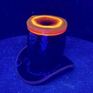 May include: A top hat shaped glass sculpture featuring a deep black body with a vibrant, glowing orange and yellow circular rim at the top. The piece exhibits a smooth, reflective surface and is illuminated under ultraviolet light, creating a high contrast.
