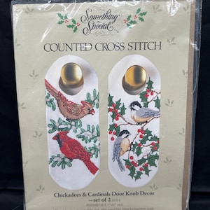 May include: A packaged counted cross-stitch kit featuring two door knob decor pieces. The designs include a red cardinal and a brown bird with green leaves and red berries. The text reads "Chickadees & Cardinals Door Knob Decor - set of 2". Finished size is 5" x 12" each.