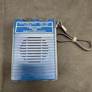 Peut inclure: Une radio météo Realistic Mini bleue avec une dragonne noire. La radio dispose d'un bouton de réglage, d'un contrôle du volume et la plage de fréquences 162,40 - 162,55 MHz est imprimée sur le devant. La radio est rectangulaire avec une grille de haut-parleur.
