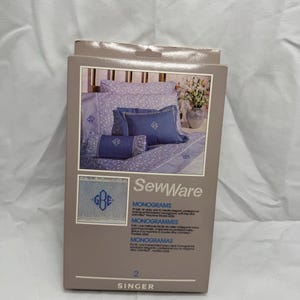 May include: A box of Singer SewWare monogramming supplies. The box is gray and features an image of a blue and white floral bed set with monogrammed pillows. The box also displays the word "MONOGRAMS" in English, French, and Spanish.