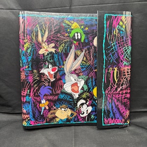 May include: A black notebook with a colorful design featuring Looney Tunes characters like Bugs Bunny, Sylvester, and the Road Runner. The cover has a neon scribble pattern in pink, yellow, and blue.