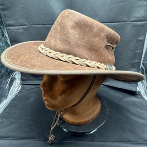 May include: A brown leather cowboy hat with a braided leather band and a brown leather chin strap.