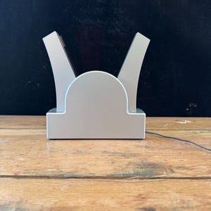 May include: A silver-colored electronic charging dock featuring a central semi-circular base with two upward-curving arms extending from the sides. The device has a smooth, matte finish and includes a thin black power cord extending from the right side.