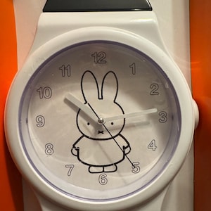 Può includere: Orologio da polso bianco con un design a coniglietto Miffy in bianco e nero sul quadrante. L'orologio ha una lunetta bianca, un cinturino nero e mostra i numeri da 1 a 12. Le lancette sono bianche.