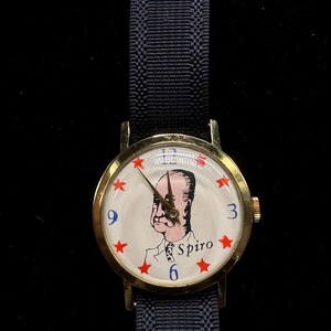 May include: A vintage wristwatch with a gold-tone case and a navy blue fabric band. The watch face features a portrait of a man, red stars, and the name "Spiro". The watch has the numbers 3, 6, and 9.