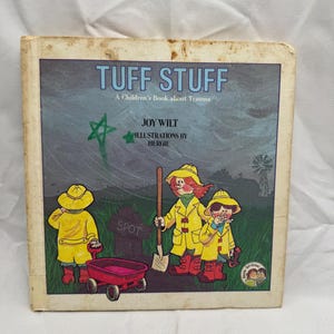 May include: A vintage children's book titled "TUFF STUFF" with illustrations by Hergie. The cover features three children in yellow raincoats and hats, with a red wagon and a green star. The book is about trauma.