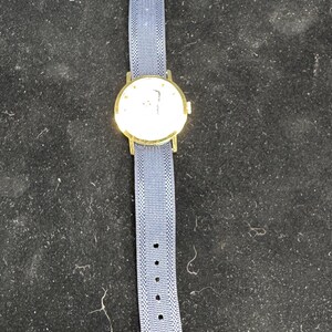 May include: A gold-toned wristwatch with a round face and a blue fabric band. The watch has a light-colored face with a simple design. The blue band has a woven texture and a gold-toned buckle.