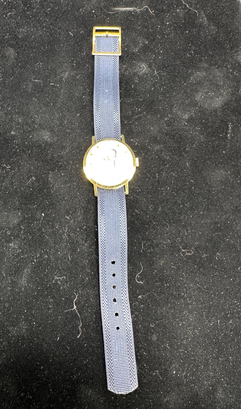 May include: A gold-toned wristwatch with a round face and a blue fabric band. The watch has a light-colored face with a simple design. The blue band has a woven texture and a gold-toned buckle.