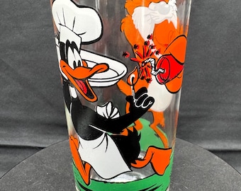 May include: Clear glass tumbler with a cartoon image of Daffy Duck in a chef's hat and apron, holding a plate. The tumbler also depicts a cartoon character with orange fur and a lit firecracker. The base of the glass has a green stripe.
