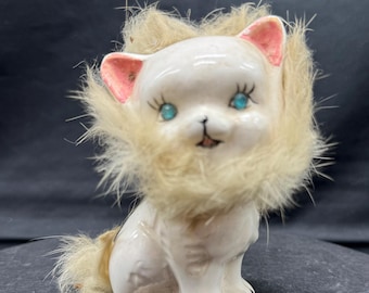 May include: A white ceramic cat figurine featuring bright blue eyes, pink-painted inner ears, and black-painted claws. The cat is adorned with a fluffy, light beige faux fur mane around its neck and a matching tuft of fur on its tail.