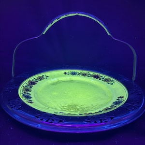 以下が含まれることがあります： 縁に花柄が施された銀製のサービングトレイ。トレイにはハンドルが付いており、金属製です。トレイはブラックライトの下で黄色く光っています。