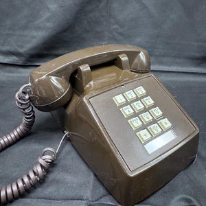 May include: A brown vintage telephone with a push-button keypad. The phone has a handset, a coiled cord, and a rectangular base. The keypad features white buttons with numbers and letters.