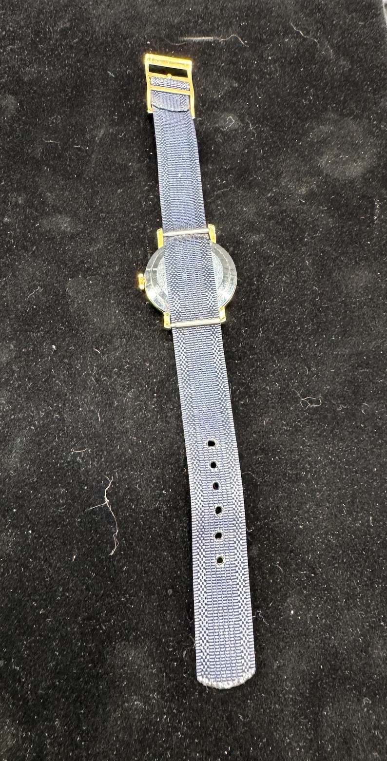 May include: A wristwatch with a blue and white checkered pattern band and a gold-colored case. The watch band has several holes for adjustment. The watch is set against a dark background.