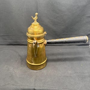 Puede incluir: Una cafetera de latón vintage con un asa negra. La olla tiene un pájaro decorativo en la tapa y detalles ornamentados. La cafetera es un diseño clásico, perfecto para preparar café.