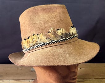 May include: A tan suede hat with a wide brim and a cylindrical crown. The hat is adorned with a band of feathers in shades of brown, black, and tan, and a braided black and white rope. The hat is displayed on a brown mannequin head.