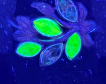May include: A ring with a floral design, featuring glowing neon green and blue accents. The ring's petals and leaves are a deep purple, creating a striking contrast against the bright green and blue. The ring is set against a dark blue background.