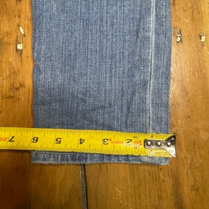 May include: Close-up of the bottom hem of a pair of blue denim jeans measured with a yellow tape measure. The tape measure reads 2, 3, 4, 5, 6, and 7 inches.