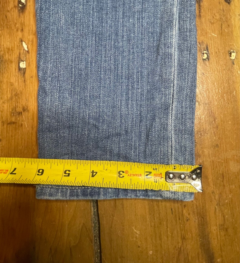 May include: Close-up of the bottom hem of a pair of blue denim jeans measured with a yellow tape measure. The tape measure reads 2, 3, 4, 5, 6, and 7 inches.