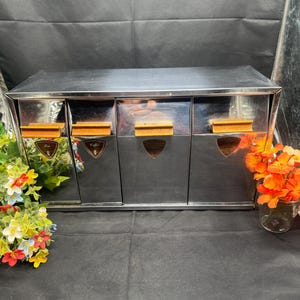 May include: A vintage chrome coffee and tea storage cabinet with three drawers. Each drawer has a wooden handle and a label. The cabinet is rectangular and has a shiny, reflective surface. Decorative flowers are on either side.