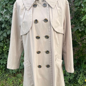 May include: A beige trench coat with a double breasted closure and a large collar. The coat has six large buttons and two pockets.