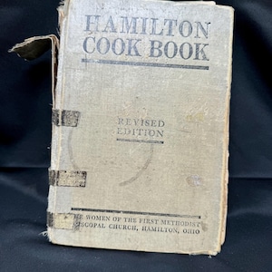 Könnte beinhalten: Ein Vintage-Kochbuch mit dem Titel "Hamilton Cook Book" und dem darunter gedruckten Text "Revised Edition". Das Buch ist in beigefarbenes Papier mit schwarzem Text gebunden und hat ein abgenutztes, gealtertes Aussehen. Der untere Teil des Umschlags lautet "The Women of the First Methodist Episcopal Church, Hamilton, Ohio."
