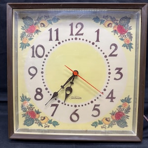 Könnte beinhalten: Eine quadratische, alte Sunbeam-Wanduhr mit braunem Rahmen. Das Zifferblatt ist gelb mit schwarzen Zahlen und Zeigern sowie einem roten Sekundenzeiger. Blumen- und Fruchtdesigns schmücken die Ecken. Die Uhr ist elektrisch.