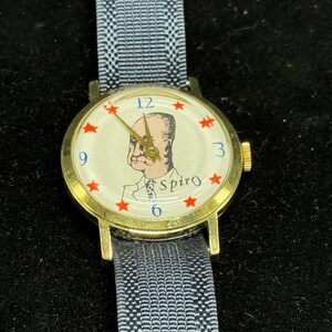 May include: A vintage wristwatch with a gold-tone case and a blue fabric band. The watch face features a portrait, red star accents, and the name "Spiro". The watch hands are gold-toned.