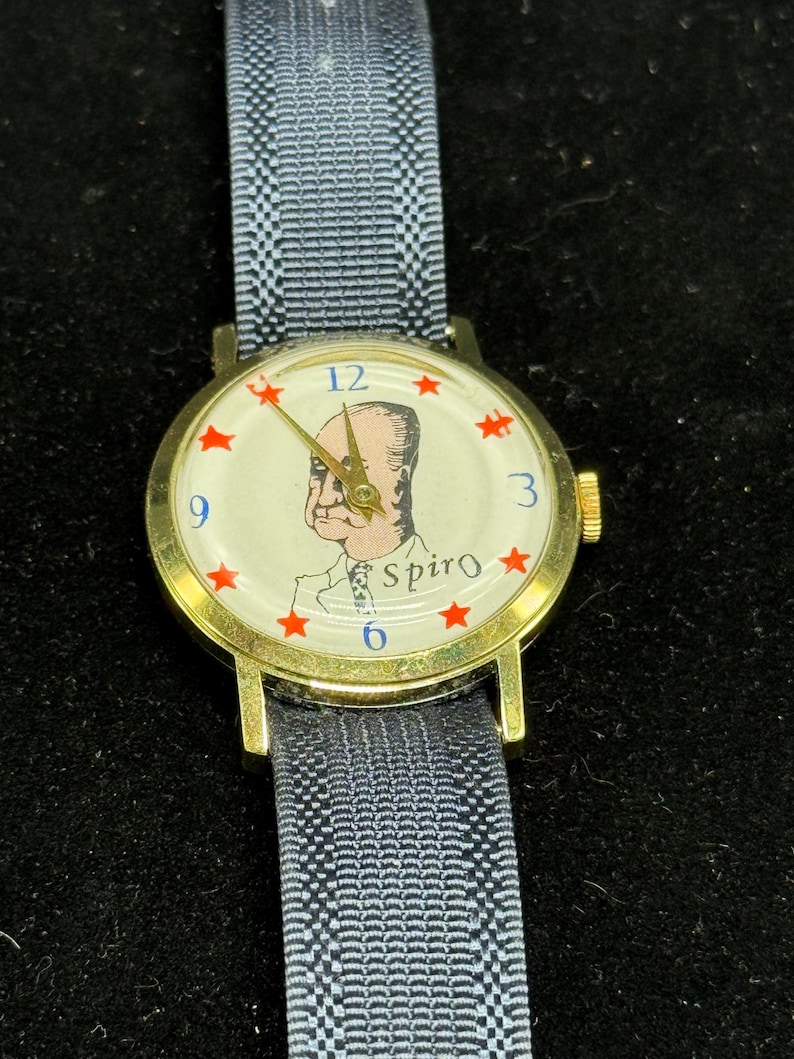 May include: A vintage wristwatch with a gold-tone case and a blue fabric band. The watch face features a portrait, red star accents, and the name "Spiro". The watch hands are gold-toned.